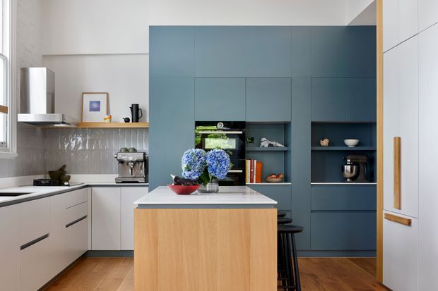 A bright contemporary kitchen with a central island featuring a light timber base and pale stone benchtop. The room has matte blue floor-to-ceiling cabinetry with integrated shelving and appliances, white cabinetry along the perimeter, a pale tiled splashback, timber flooring, and black stools. Natural light enters from the window at left, creating a clean yet warm atmosphere.