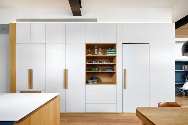 A bright contemporary kitchen centred on a full-height wall of smooth white cabinetry, with warm timber handles and a timber open display niche containing crockery and glassware. A white island with timber sides is visible in the foreground, along with a timber dining table and leather seating. The room has timber flooring, a dark exposed ceiling beam, integrated ventilation, and partial views toward an adjacent living area.