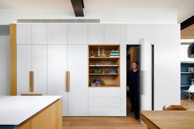 Bright contemporary kitchen interior with a large white island in the foreground, extensive flat-panel white cabinetry, warm timber side panels and integrated vertical handles, and an open timber-lined display shelving niche. A concealed doorway is integrated into the cabinetry, with timber flooring and a dining table visible at the right. The room has a clean, restrained fit-out with a dark exposed ceiling beam and partial black-framed glazing.