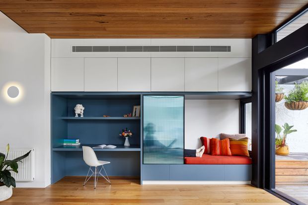 Contemporary built-in study and window seat within a compact open room. White overhead cabinetry spans the wall above a deep blue shelving and desk nook, with a frosted-glass cabinet door and a red-orange upholstered bench seat. Warm timber ceiling and flooring contrast with dark-framed full-height glazing opening toward an outdoor deck or courtyard. Clean lines, integrated joinery and abundant natural light define the space.