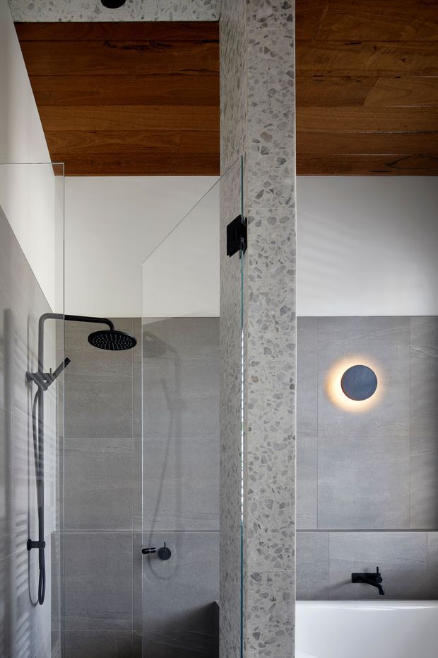 A contemporary bathroom featuring a walk-in glass shower and a partially visible freestanding bathtub. Large grey wall tiles line the wet areas, while a terrazzo-clad dividing column adds texture. The ceiling is finished in warm timber boards, contrasting with white walls and matte black shower and tapware. A circular wall light provides a soft focal accent.