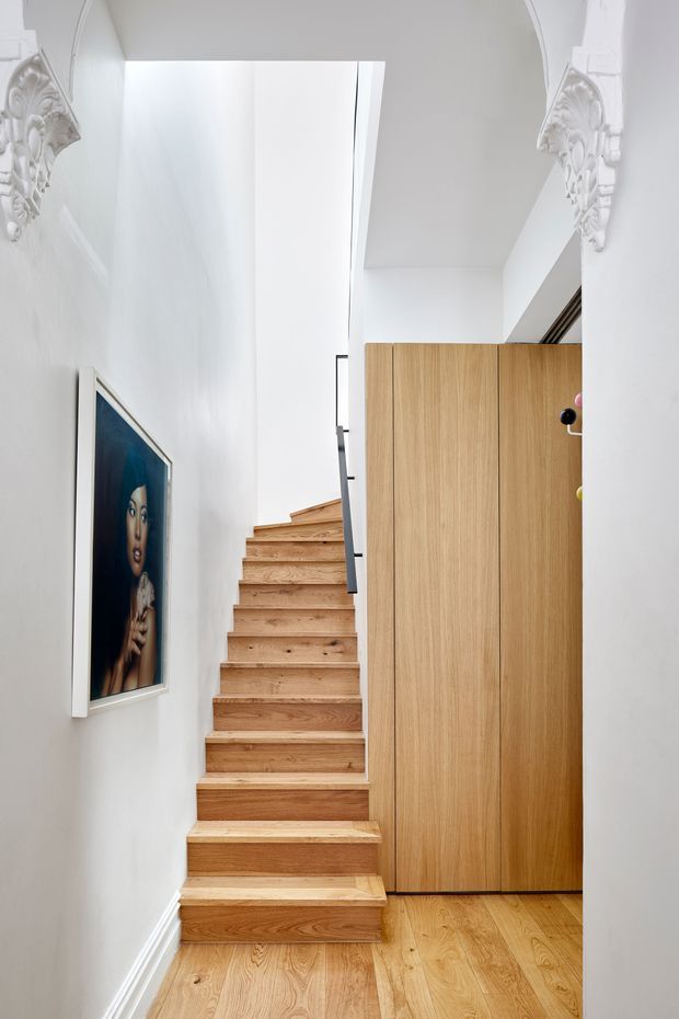 A narrow, bright residential stairway with warm timber stairs and matching timber flooring. White walls feature ornate decorative plaster corbels near the ceiling, while a tall flush timber storage or wardrobe joinery unit sits beside the stairs. A framed artwork hangs on the wall, and a skylit or double-height opening brings in abundant natural light.