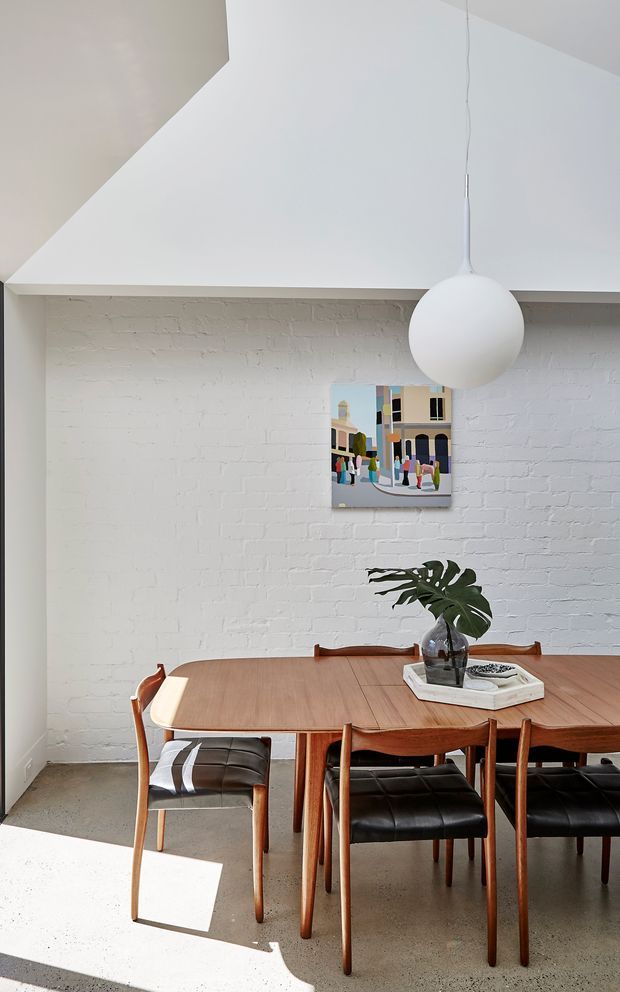 Bright dining area with a mid-century-style timber dining table and matching wooden chairs with black leather seats, set against a white-painted exposed brick wall. A large white globe pendant hangs above the table, with a colourful artwork and simple tropical leaf arrangement adding visual interest. Sunlight creates strong geometric patches across the polished concrete floor.