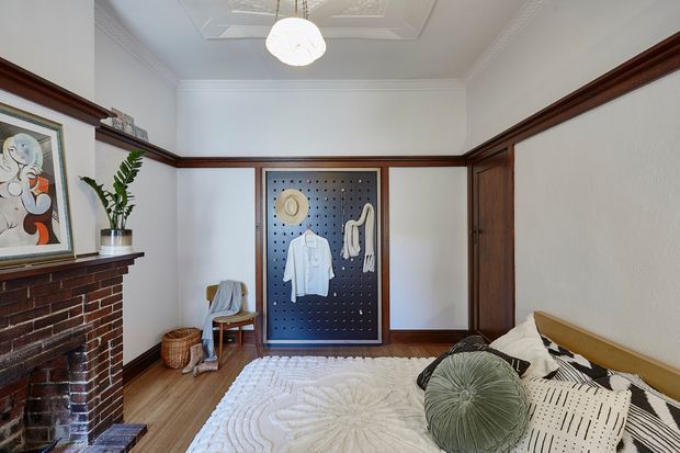A traditionally detailed bedroom with white walls, dark timber dado and trim, a red brick fireplace, timber flooring, and ornate pressed ceiling panels. The room features a bed with neutral patterned bedding, a decorative round cushion, a framed artwork, a freestanding clothing display board, and a small chair and basket. The overall character blends heritage architectural detailing with eclectic contemporary furnishings.