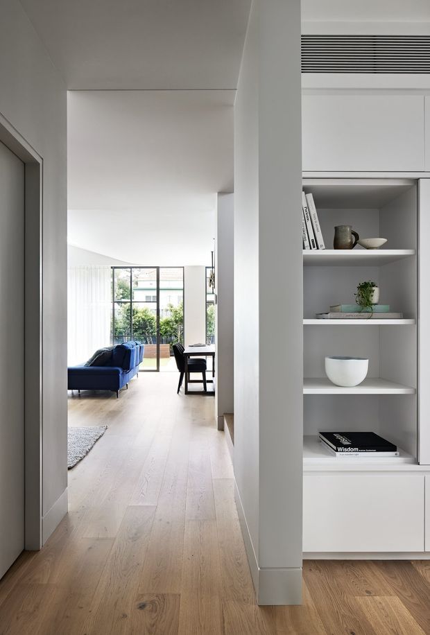 A bright, minimalist residential interior viewed from a hallway, looking toward an open living and dining area. Pale timber flooring runs through the space, with a deep blue sofa, black-framed glazed doors, and abundant natural light. On the right, a full-height white built-in shelving and storage unit displays books and ceramics, with a linear ventilation grille above. The palette is restrained and contemporary, with clean white walls and subtle architectural detailing.