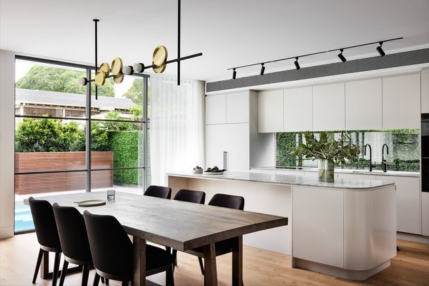 A bright, contemporary open-plan dining and kitchen interior. A large dark timber dining table with black upholstered chairs occupies the foreground, while a streamlined white kitchen with an island, integrated cabinetry, and a mirrored splashback sits behind it. Full-height glazing opens toward a landscaped outdoor area, and a sculptural brass-and-black pendant light hangs above the dining table. Track lighting and a linear ceiling ventilation grille add modern architectural detailing.