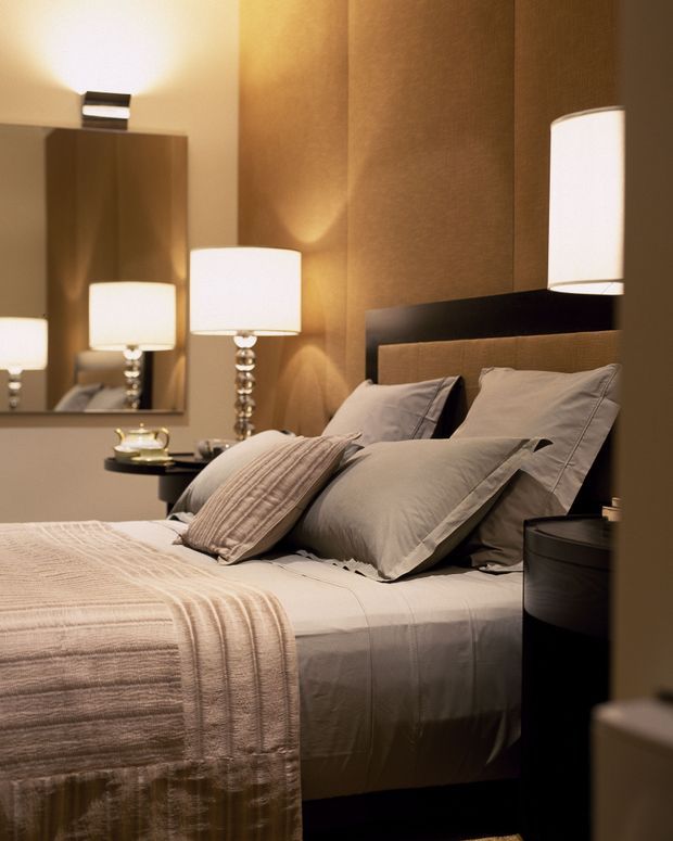 A warmly lit bedroom featuring a neatly made bed with layered beige and taupe bedding, a dark upholstered headboard, bedside tables, contemporary table lamps, and a large wall mirror. Textured warm brown wall finishes and soft neutral furnishings create an intimate, refined atmosphere.