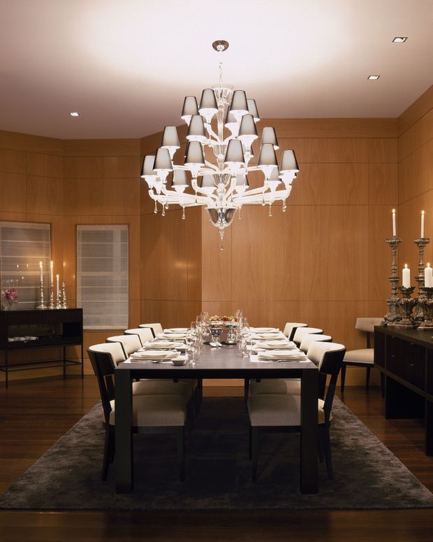 Formal dining room with a long dark dining table set for a meal, surrounded by upholstered dining chairs. The room features warm timber wall panelling, dark timber flooring, a large ornate crystal chandelier, candlelit candelabras, a sideboard, and low contemporary lighting. The atmosphere is refined and dramatic.