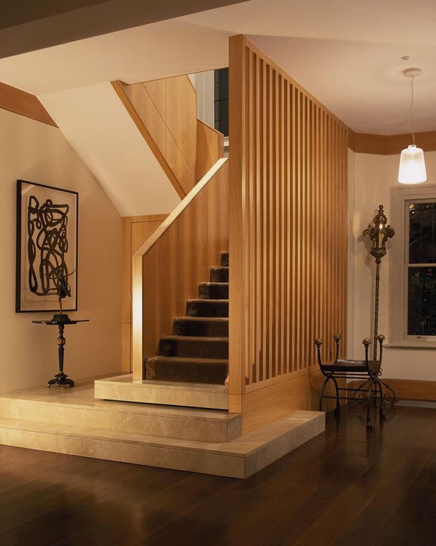 Warm, refined interior view of a timber-lined stairway and landing. A dark carpeted staircase rises beside a full-height vertical timber slat screen, with matching timber wall panelling and cabinetry. Pale stone-clad stair platforms contrast with dark polished timber flooring. Traditional decorative furnishings, framed artwork, a pendant light and a window complete the elegant composition.