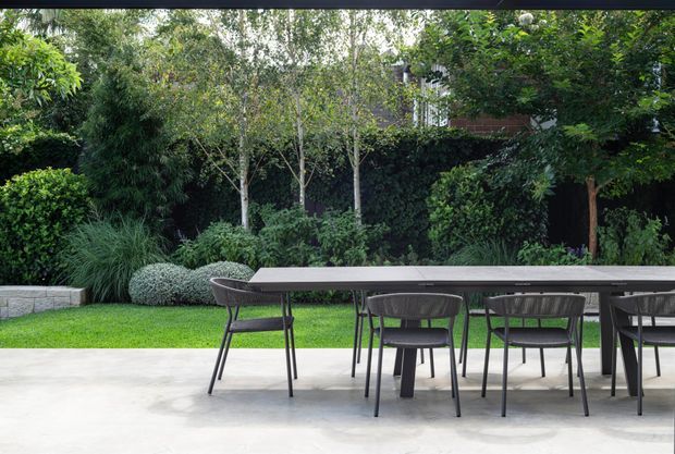A landscaped outdoor dining terrace with a long dark rectangular dining table and matching chairs on a pale paved surface. A neatly manicured lawn, layered shrubs, mature trees, and dense hedging form a private garden backdrop, with part of a residential building visible behind the planting.