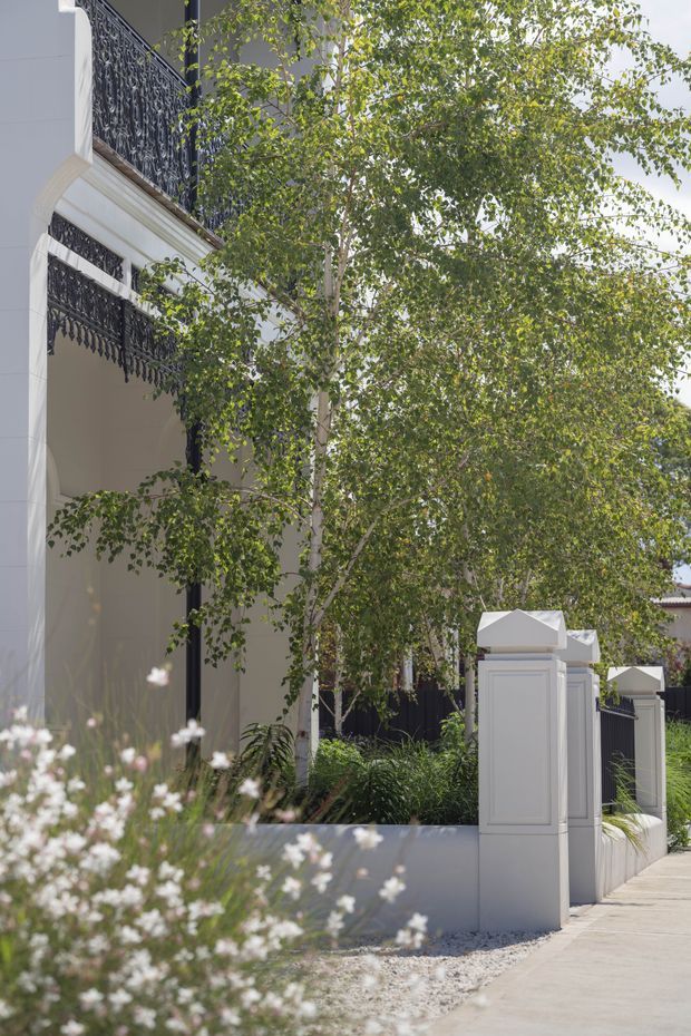 A refined residential facade viewed from the street, featuring white rendered walls, ornate black wrought-iron balcony balustrades, decorative white gate pillars, and layered garden planting with slender trees and soft flowering plants. The image has a bright, elegant suburban character.
