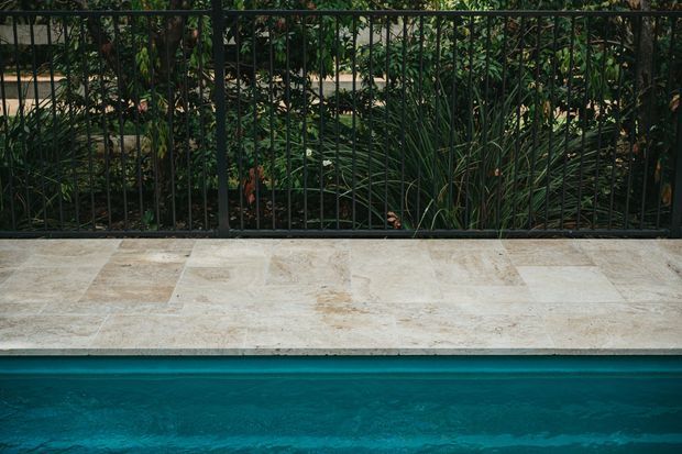Close-up view of a turquoise swimming pool bordered by a light beige stone-tiled coping or terrace edge, with a black metal railing and dense planted garden beyond.