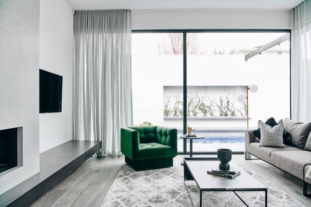 Bright contemporary living room with floor-to-ceiling glazing overlooking a courtyard pool. The room features a white textured fireplace wall with wall-mounted television, a long dark hearth, sheer grey curtains, a deep green tufted armchair, a large grey sofa, and a black-framed rectangular coffee table on a patterned rug.