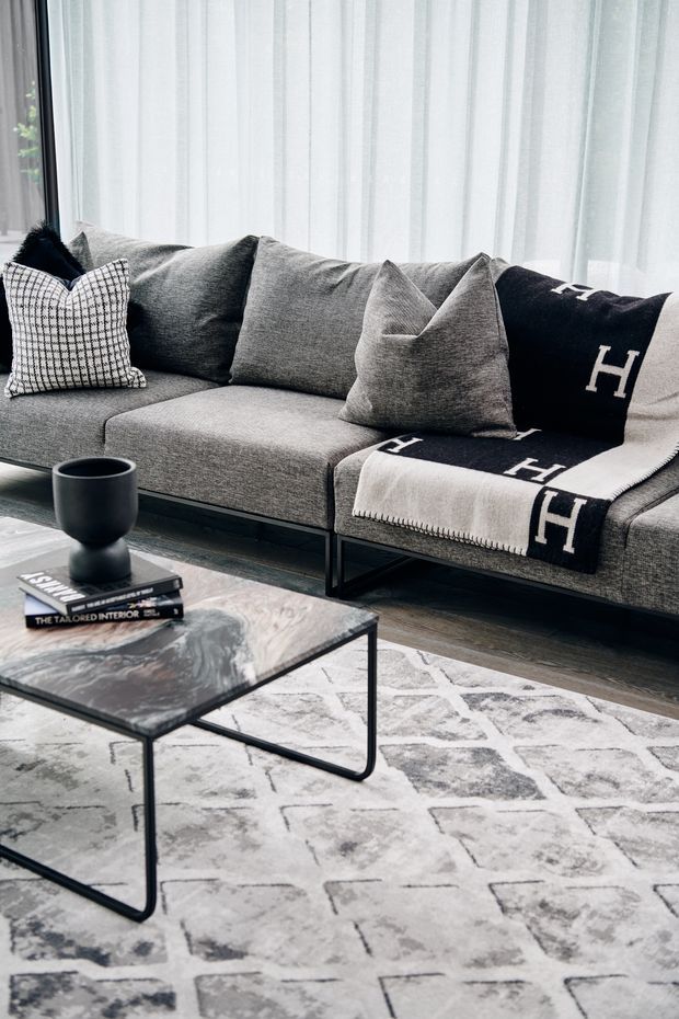 A styled contemporary living room featuring a large grey upholstered sectional sofa with layered cushions and a black-and-cream monogram throw. Sheer floor-to-ceiling curtains provide soft natural light. In front of the sofa is a slim black metal coffee table with a dark marbled stone-like top, set over a pale abstract patterned rug. The palette is monochromatic grey, black and white with refined, luxe accents.