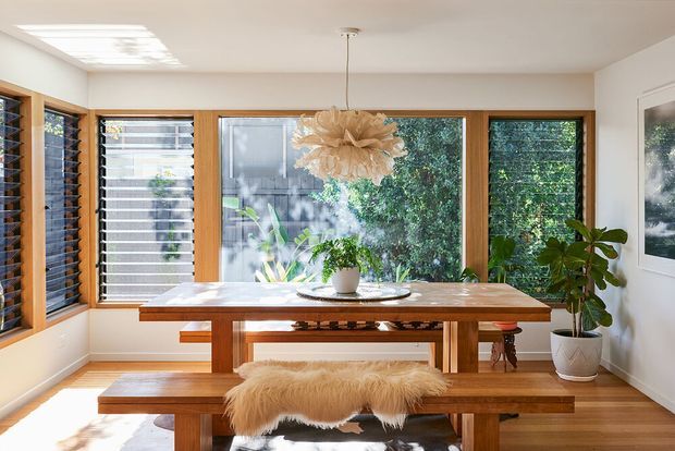 Bright dining area with a substantial timber dining table and matching bench, surrounded by large timber-framed windows and louvred glazing overlooking greenery. A sculptural pendant light hangs above the table, while potted plants and a sheepskin throw add natural texture. The room has white walls, warm timber flooring and joinery, and abundant daylight.