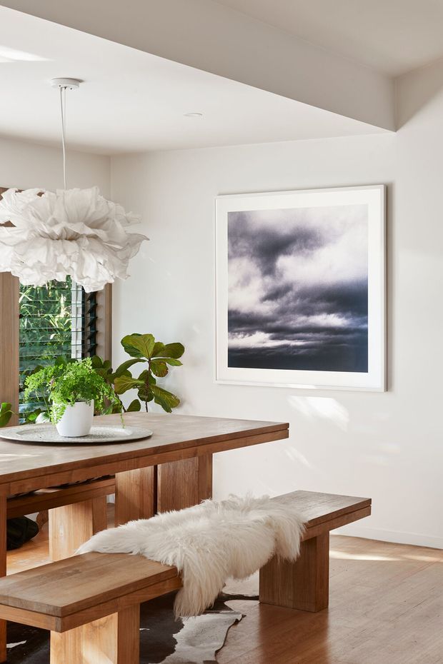 Bright, warm dining area with a substantial solid timber dining table and matching benches. The room has white walls and ceiling, pale timber flooring, a large white sculptural pendant, potted greenery, a framed monochrome cloud photograph, and a louvred window bringing in sunlight. A soft white sheepskin is draped over one bench, creating a natural, relaxed atmosphere.