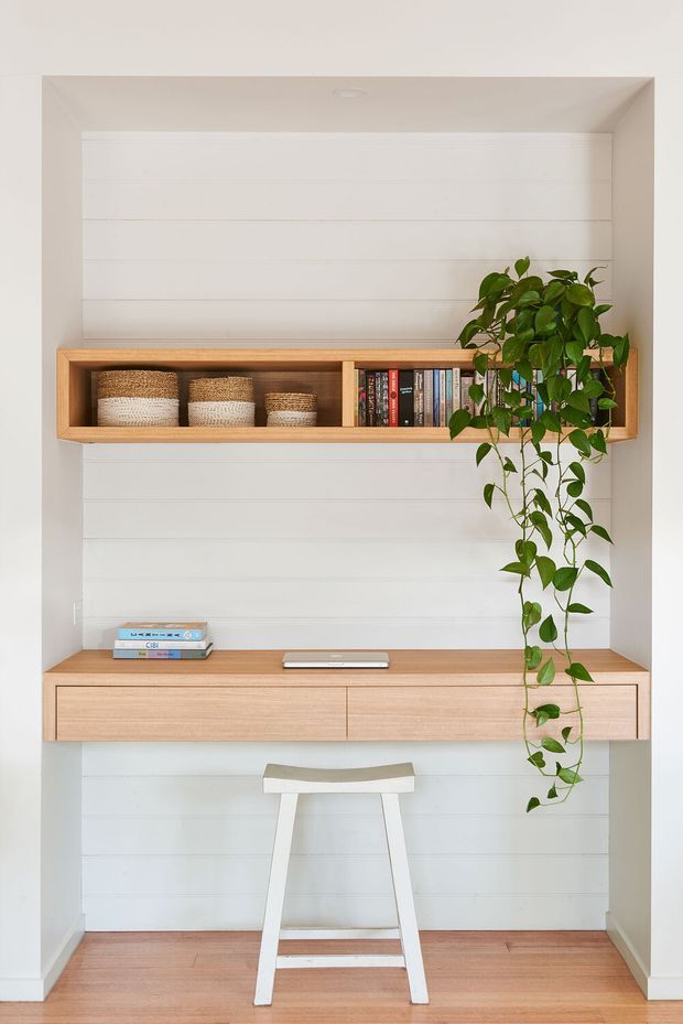 A bright, compact built-in home office nook with white horizontal wall panelling, a floating light timber desk with drawers, and a matching open wall shelf containing books and woven baskets. A trailing indoor plant adds greenery, while a simple white stool sits beneath the desk. The space has clean lines, warm timber accents, and a minimal, contemporary feel.