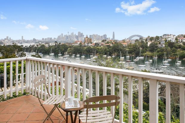 A sunny residential balcony or terrace overlooking a marina and leafy waterfront, with white-painted vertical balustrades, terracotta tiled flooring, and simple outdoor seating. A dense urban skyline and a large arched bridge are visible across the water in the distance.