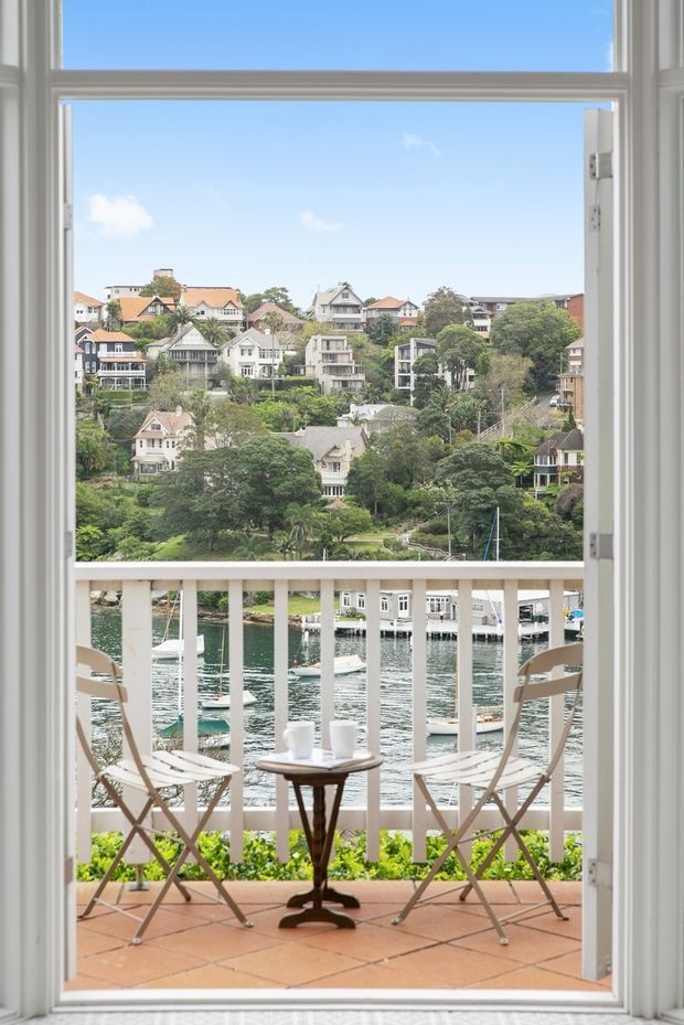 View from inside through an open white-framed glazed door onto a small tiled balcony with white balustrade, two folding chairs and a small table. Beyond the balcony is a leafy waterfront setting with boats, calm water, and densely built hillside residences. The image is bright and airy with a coastal, relaxed character.