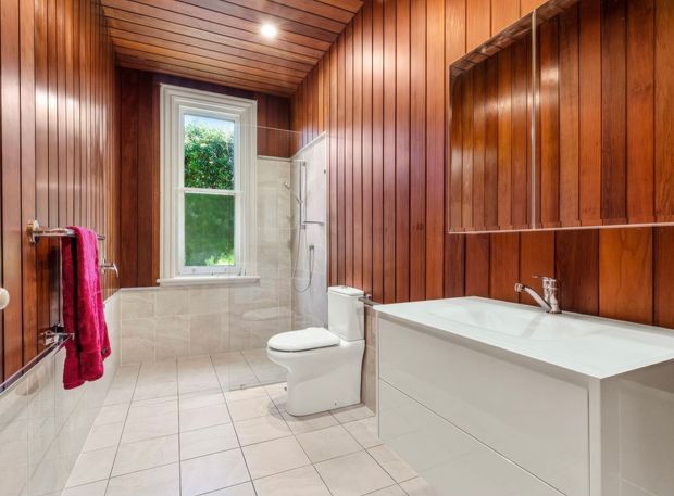A warm, narrow bathroom featuring extensive mid-tone timber wall and ceiling panelling, light tiled flooring and shower surround, a freestanding-style toilet, and a white built-in vanity with integrated basin. A window provides natural light, while an open timber medicine cabinet and towel rail are visible.