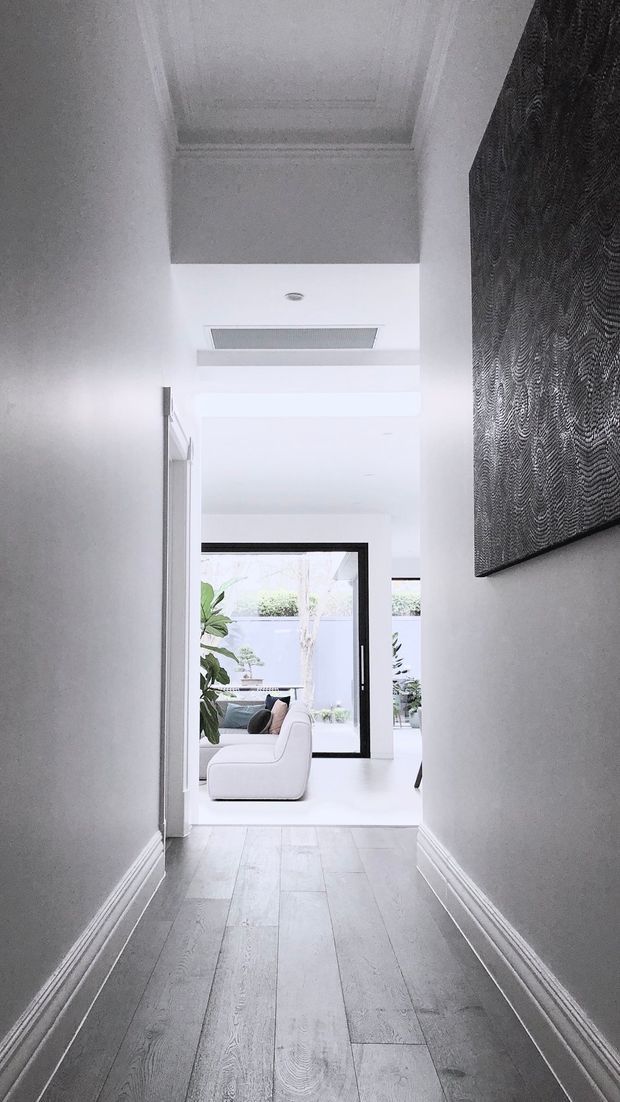 A bright, narrow residential hallway with white painted walls, decorative skirting boards, pale timber flooring, and a high coffered ceiling. The corridor opens into a light-filled living area with a white curved sofa, indoor plants, and black-framed glazing leading to an outdoor courtyard. A large monochrome textured artwork forms a feature on the right wall, while a linear ceiling air vent and recessed lighting add contemporary detailing.
