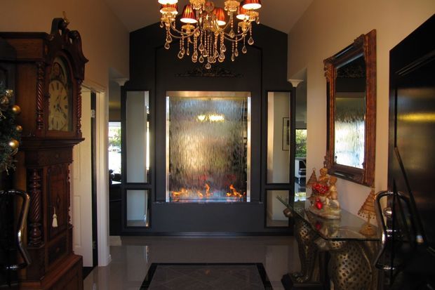 A dramatic, richly styled entry or living area with a dark feature wall centred on a glass-fronted fireplace, flanked by narrow decorative windows. The room includes an ornate wood grandfather clock, large carved-framed mirror, glass console table, and an elaborate crystal chandelier. Glossy flooring and black, bronze, and warm wood finishes create a formal, luxurious atmosphere.