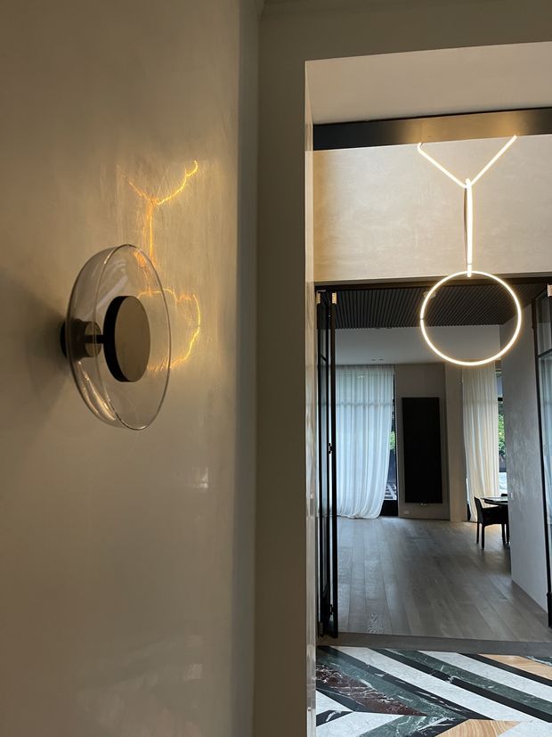 A contemporary interior hallway viewed through a doorway, featuring a sculptural wall sconce with a clear circular glass shade and warm integrated lighting. A large geometric pendant light hangs beyond the doorway, illuminating a refined open room with pale timber flooring, sheer curtains, dark architectural details, and a bold geometric stone or tile floor pattern in the foreground.