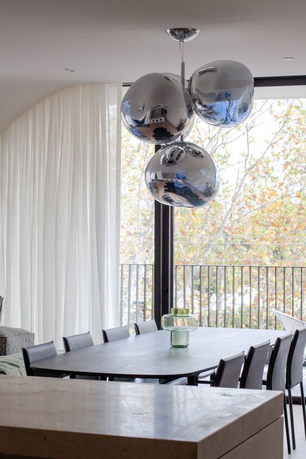 A contemporary dining area with a dark oval dining table and matching upholstered chairs, positioned beside expansive floor-to-ceiling glazing opening to a balcony. A sculptural cluster pendant with reflective chrome globes hangs above the table. Sheer white curtains soften the large windows, while a pale stone kitchen island occupies the foreground. The space is bright, refined, and connected to leafy outdoor views.