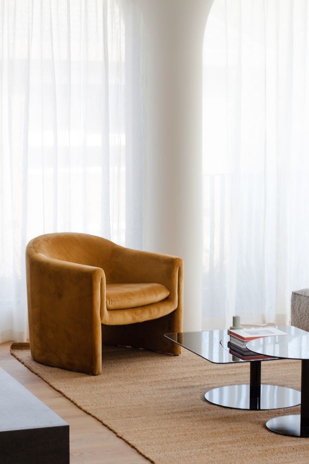 Bright, refined living room corner with a sculptural caramel leather tub chair positioned beside a low glass-top coffee table with a black pedestal base. A natural woven rug, pale timber flooring, sheer full-height curtains, and a curved light-coloured wall create a soft, airy setting. Stacked books and a small cylindrical object sit on the table, with part of a dark low cabinet visible in the foreground.