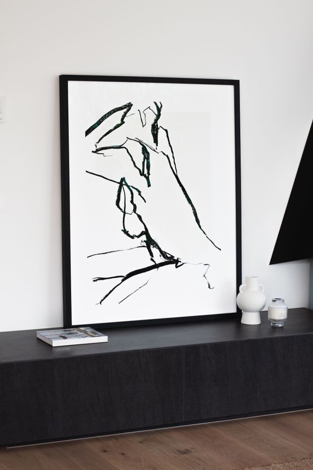 A minimalist interior vignette featuring a large black-framed abstract line-art print leaning against a white wall above a long dark built-in sideboard. Small sculptural ceramic objects, a candle, and a book sit on the sideboard, with warm timber flooring below and part of a black angular floor lamp visible at right.