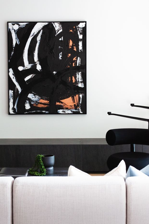 Contemporary living room viewed from behind a light upholstered sofa. A dramatic black, white, and burnt-orange abstract artwork hangs above a dark timber built-in cabinet, with a black sculptural lounge chair and small potted plant adding contrast. The space has a restrained, monochromatic palette with bold artwork and clean-lined furnishings.