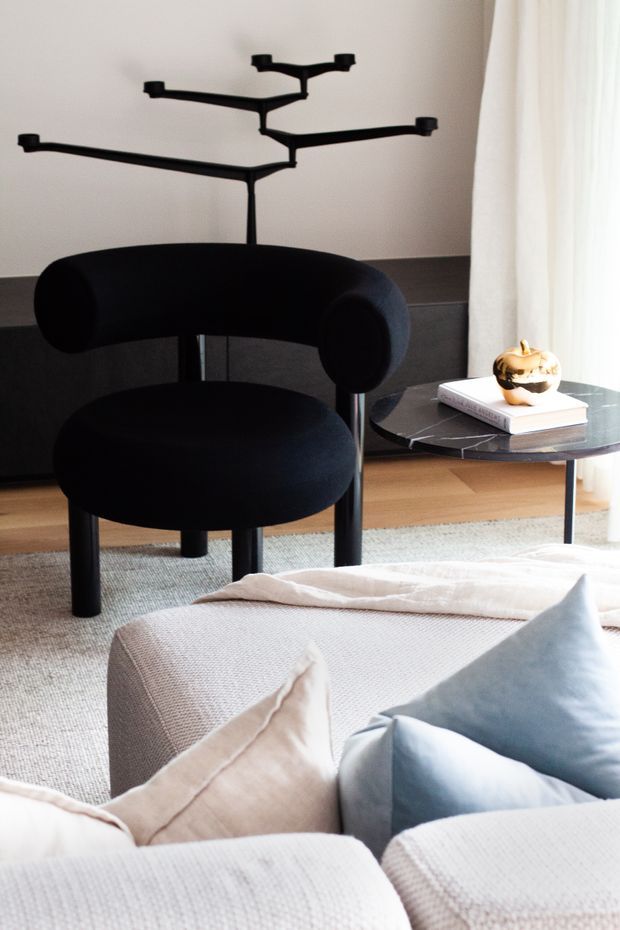 A refined contemporary living room vignette featuring a sculptural black lounge chair, a black branching pendant light, a low dark marble-topped side table, and a pale upholstered sofa with soft blue and cream cushions. Dark wall panelling contrasts with light flooring, sheer curtains, and warm natural daylight.