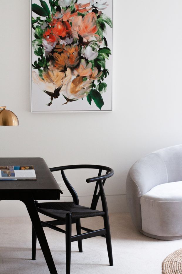 A refined interior vignette featuring a black timber desk, woven-seat black armchair, curved light upholstered lounge chair, brass desk lamp, pale carpet, and a large framed floral artwork on a soft white wall. The room has a restrained, contemporary palette with warm artistic accents.