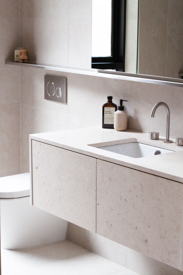 A bright, minimalist contemporary bathroom featuring a wall-hung pale stone-look vanity with integrated benchtop and inset basin, brushed metal tapware, large-format light beige wall tiles, a frameless mirror, and a concealed cistern toilet. A small window provides natural light.