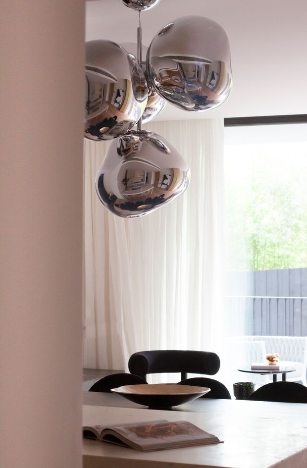 A refined contemporary dining area with a white dining table, black upholstered dining chairs, and a sculptural cluster pendant light with reflective chrome spherical shades. Sheer curtains cover large glazing, creating a bright connection to an outdoor terrace or garden. A shallow decorative bowl and open book sit on the table.