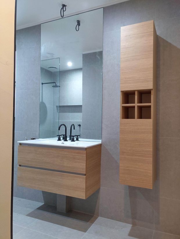 Contemporary bathroom vanity with a floating timber-look double-drawer cabinet, white integrated basin, and black curved tapware. Large mirrored wall panels reflect a glass shower enclosure, while matching timber-look wall cabinetry provides open storage niches. Large grey wall and floor tiles create a clean, neutral finish.
