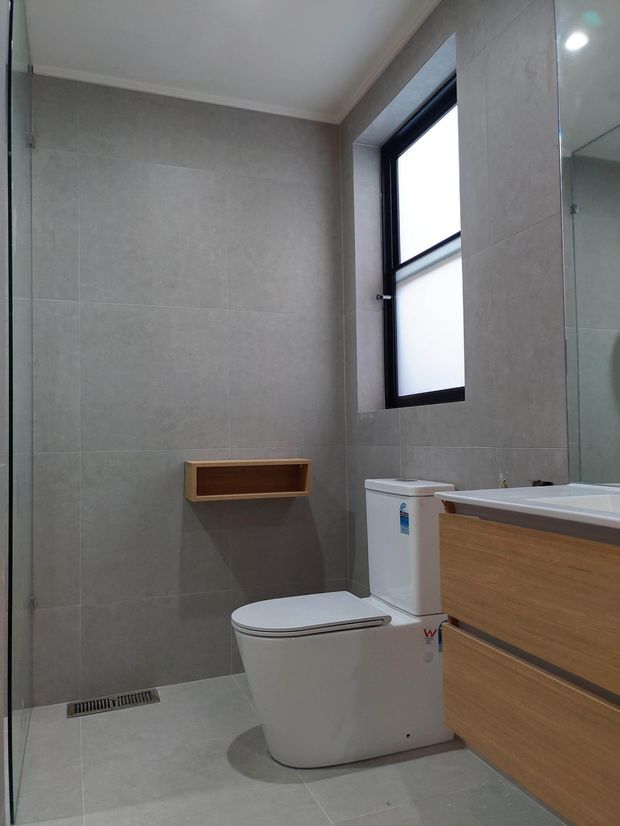 Contemporary bathroom with large-format grey wall and floor tiles, a wall-mounted timber niche shelf, white toilet, timber vanity cabinetry, frameless glass shower enclosure, and a black-framed frosted window. The room has a clean, understated finish with neutral grey and warm wood tones.