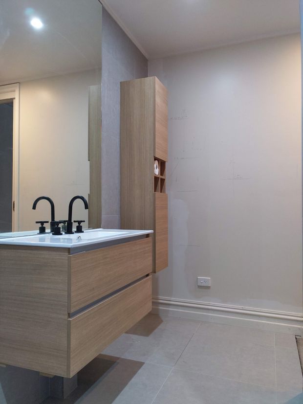 A contemporary bathroom vanity with a white integrated basin, twin black tap sets, large mirror, and light wood-grain floating cabinetry. Grey floor-to-ceiling wall tiles, pale painted walls, recessed ceiling lighting, and a narrow matching storage tower complete the space.
