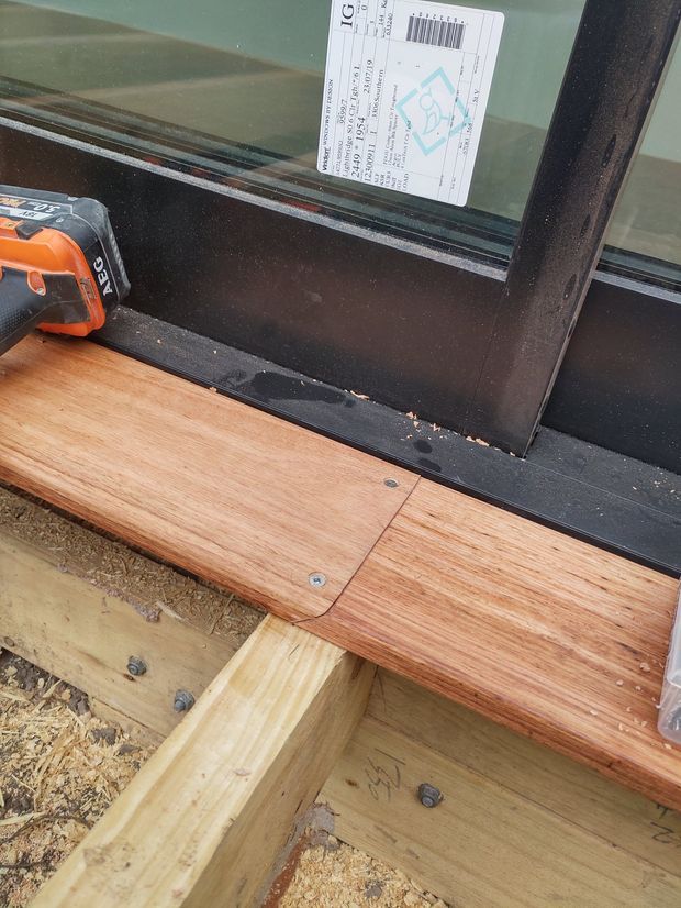 Close-up construction detail showing timber decking or flooring boards being installed along a black-framed glazed door or window. Exposed timber joists, screws, sawdust, and a cordless drill are visible, with a small construction label attached to the glazing.
