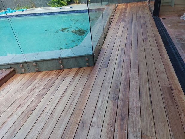 A close-up exterior view of a timber deck surrounding a swimming pool. The pool has clear glass safety balustrading with dark metal fixings, while the deck uses long, narrow warm-toned timber boards. A dark-framed glazed opening is visible at the right edge.