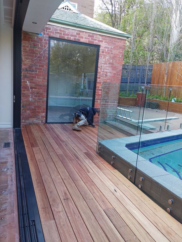 A residential outdoor deck beside a swimming pool, viewed from a covered area. The deck is finished in warm timber boards and bordered by frameless glass pool fencing. A red brick pavilion with a large glazed door and green metal roof sits at the far end, with timber and dark screening fences beyond.
