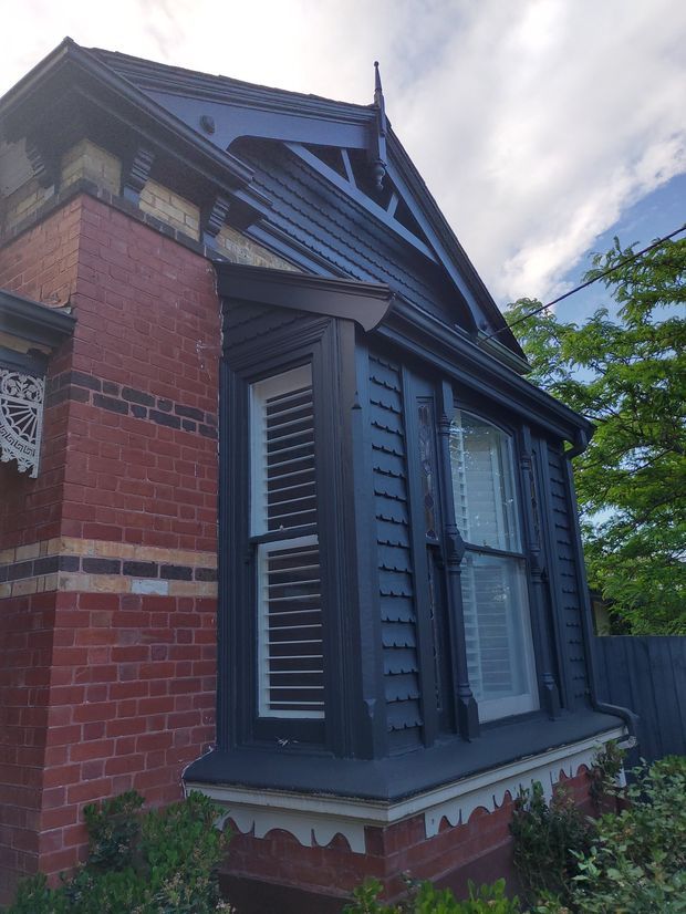 Close-up exterior view of a heritage brick residence with ornate Victorian-era detailing. The facade combines red brickwork, dark painted timber weatherboard cladding, decorative gable trim, moulded cornices, louvred shutters, and a projecting bay window with white blinds. Mature greenery is visible alongside the building.
