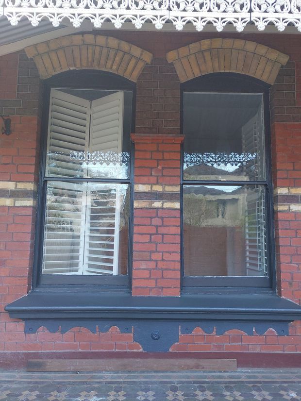 Close-up exterior detail of a heritage brick facade with two arched window openings, dark painted timber frames, white plantation shutters, decorative brick voussoirs, ornate white cast-iron lacework above, and a patterned encaustic tile pavement below.