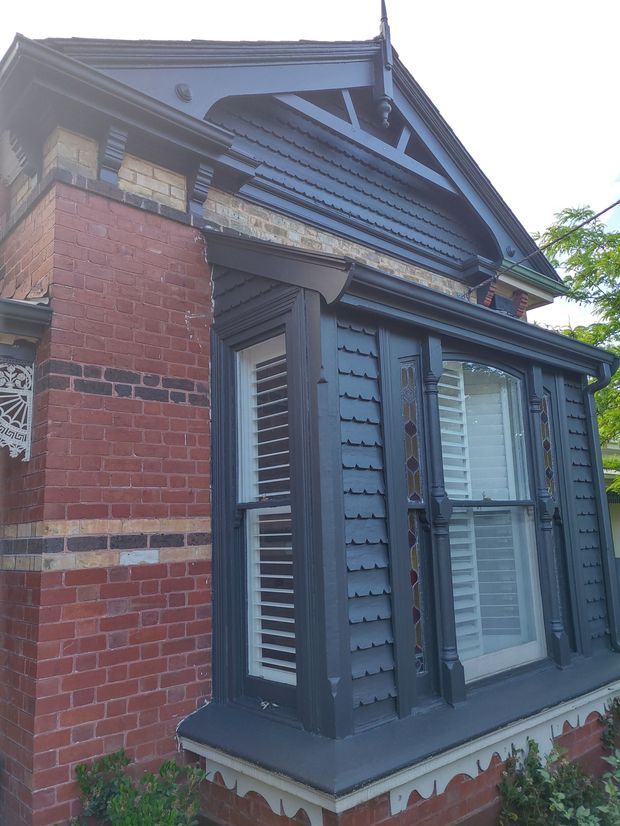 Close-up exterior view of a heritage brick house facade featuring an ornate projecting bay window, dark painted timber weatherboard cladding, decorative timber trim, louvred shutters, leadlight side panels, and a steep gabled roof with patterned bargeboard detailing. The facade combines red and cream brickwork with charcoal-grey painted timber elements.