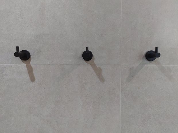 Close-up of a minimalist tiled wall with three matte black wall-mounted bathroom fixtures or tap controls arranged across large light grey tiles. Strong directional lighting creates long shadows, emphasizing the clean geometric composition.