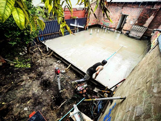 An outdoor construction site showing a worker finishing a freshly poured concrete slab in a confined courtyard or backyard area. The scene includes temporary formwork, construction tools, exposed brick and rendered boundary walls, corrugated metal fencing, and dense vegetation.