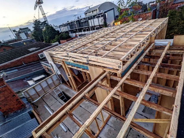 Elevated view of a timber-framed building under construction, showing exposed wall and roof framing, temporary structural supports, and adjoining urban rooftops. A construction crane and neighbouring buildings are visible in the background.