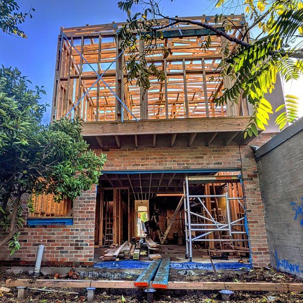 A two-storey residential building under construction, viewed from the exterior. The lower level has exposed red brick walls and an open garage or entry opening, while the upper level shows a timber structural frame with temporary bracing, scaffolding, and exposed floor and roof framing. The surrounding trees and adjacent masonry wall indicate a narrow urban or suburban site.
