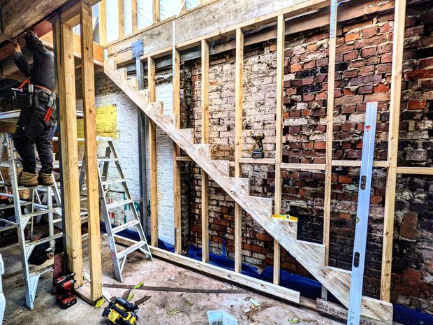 An unfinished interior renovation under construction, showing exposed timber wall and stair framing, reclaimed brick masonry, construction tools, ladders, and a worker installing structural framing.