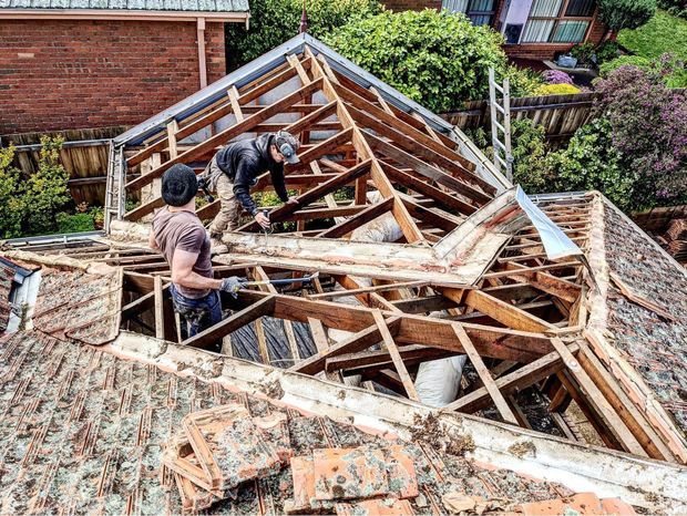 Aerial view of a residential roof undergoing major demolition or reconstruction. Two workers are standing among exposed timber rafters and roof framing, with removed and weathered terracotta roof tiles in the foreground. Brick neighbouring structures, timber fencing, and established garden vegetation are visible around the worksite.